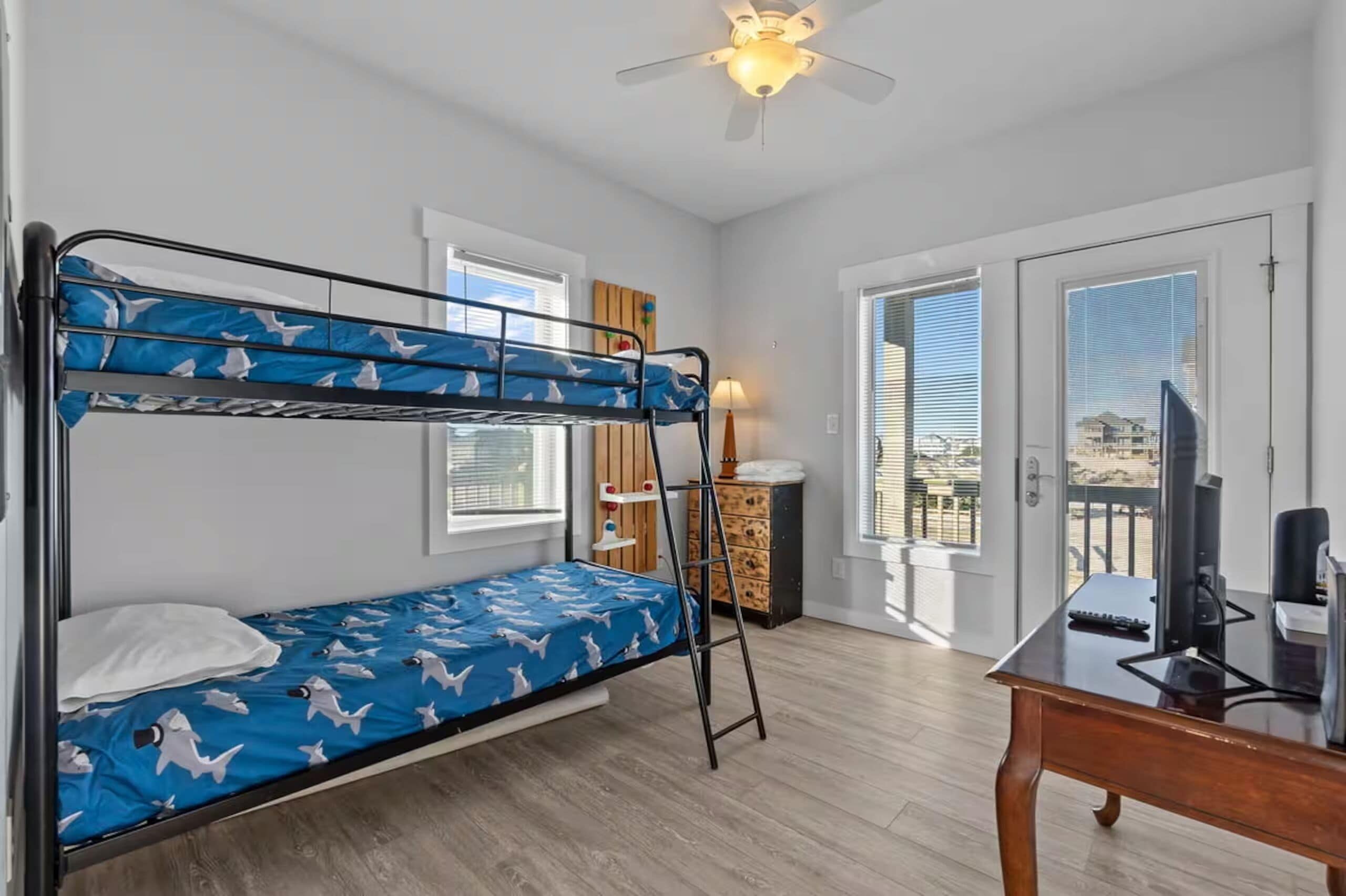 A room with a bunk bed, a desk, a chair, a television, and a window with blinds.