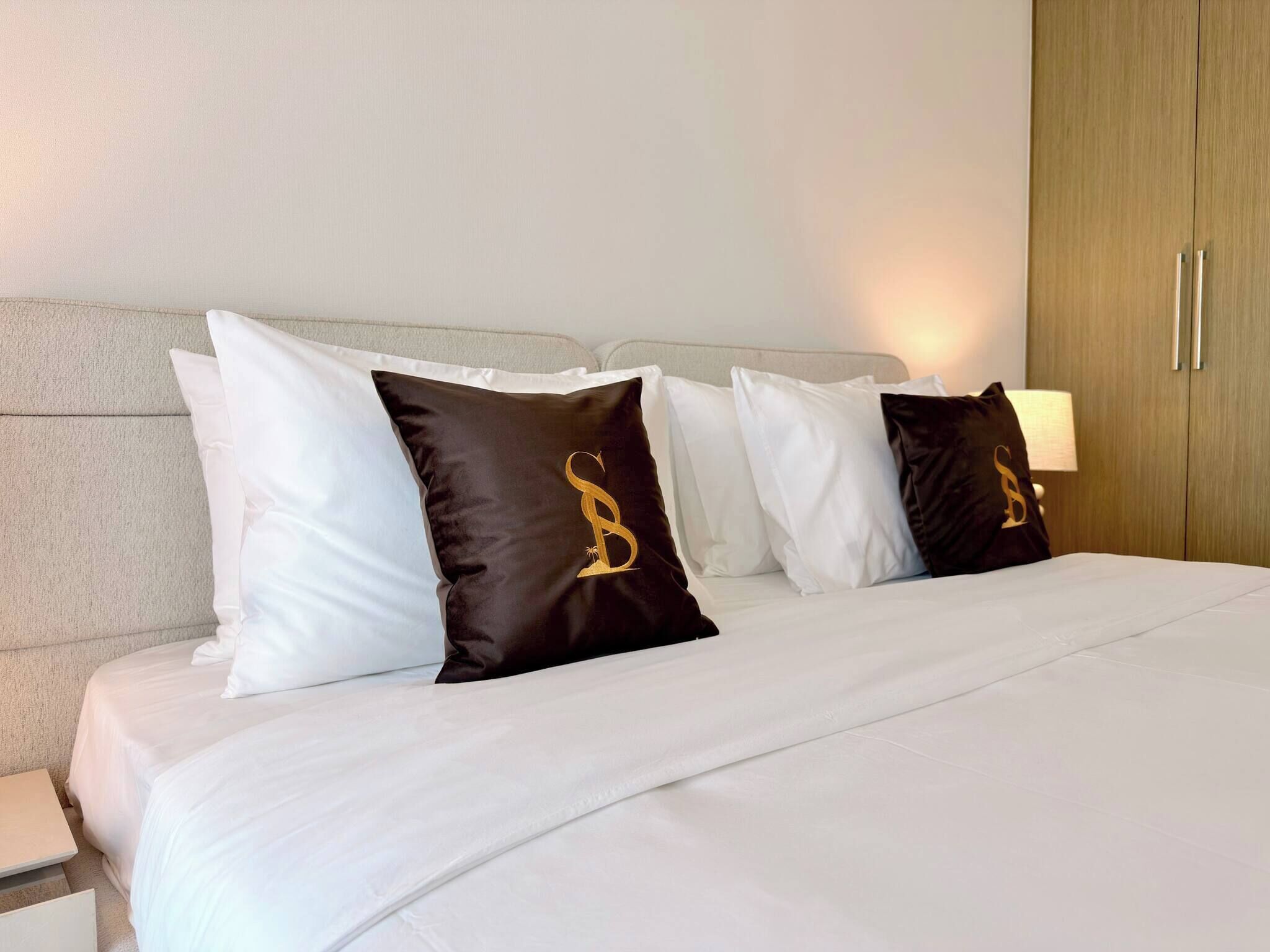 Een net opgemaakt bed met witte lakens en twee zwarte kussens met een gouden embleem.