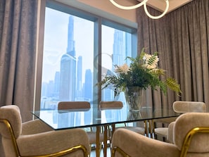 A modern dining area with a glass table, four chairs, a vase with flowers, and a cityscape view through large windows. - Burj Khalifa View Opposite to Dubai Mall Luxury 2B (Dubai)