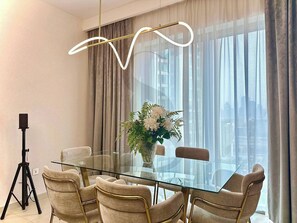 Ein moderner Essbereich mit einem Glastisch, beige gepolsterten Stühlen, einer dekorativen Vase mit weißen Blumen und einer auf einem Stativ befestigten Kamera auf der linken Seite.