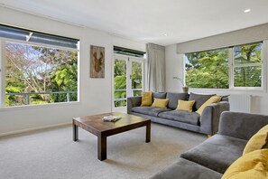 Ein modernes Wohnzimmer mit einem grauen Sofa, gelben Kissen, einem Holztisch und großen Fenstern mit Blick auf Grünflächen.