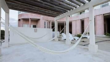 A covered outdoor area with a hammock, white lounge chairs, and a building with pink walls and a concrete floor.