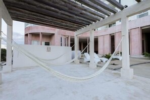 Un espace extérieur abrité comprenant un hamac, des chaises longues blanches et un bâtiment aux murs roses et au sol en béton.