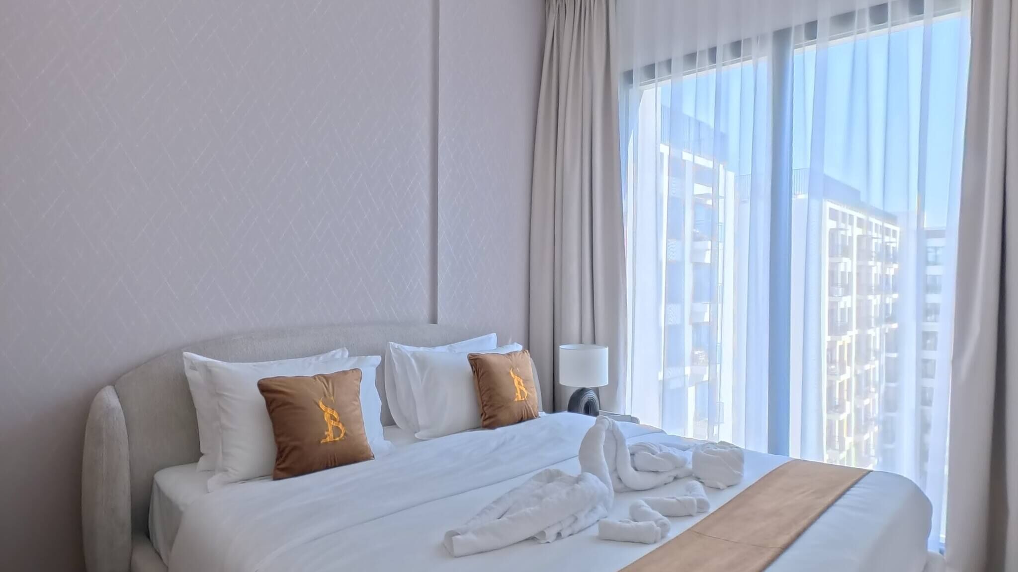 Een hotelkamer met een groot bed, witte beddengoed, een houten hoofdeinde, een nachtkastje met een lamp en uitzicht op gebouwen door dunne gordijnen.