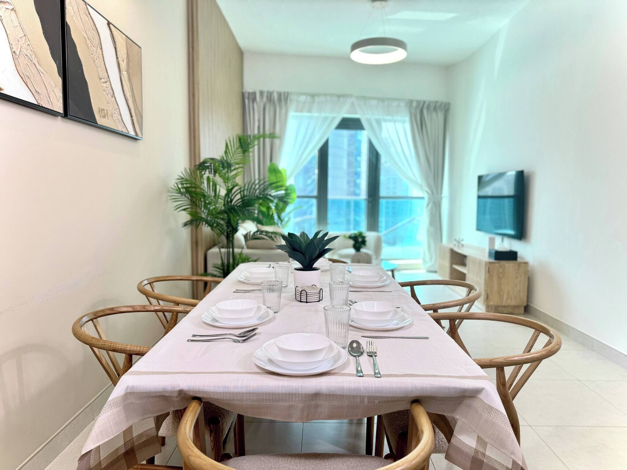 A dining area with a long table set for four, surrounded by wicker chairs, with a potted plant and a television on a stand in the background.