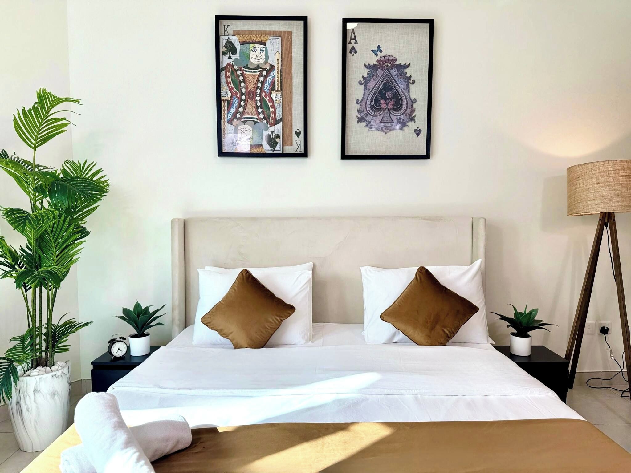 Ein ordentlich bezogenes Bett mit zwei Kissen, ein Nachttisch mit einer Uhr und einer Pflanze sowie zwei gerahmte Kunstwerke an der Wand.
