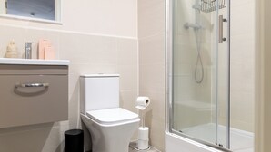 Casa de banho com sanita, um contentor de lixo, cabine de duche e suporte para papel higiénico.