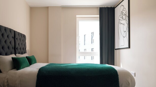 Quarto de hotel com cama, cabeceira, manta verde, obra de arte emoldurada e vista de edifícios pela janela.
