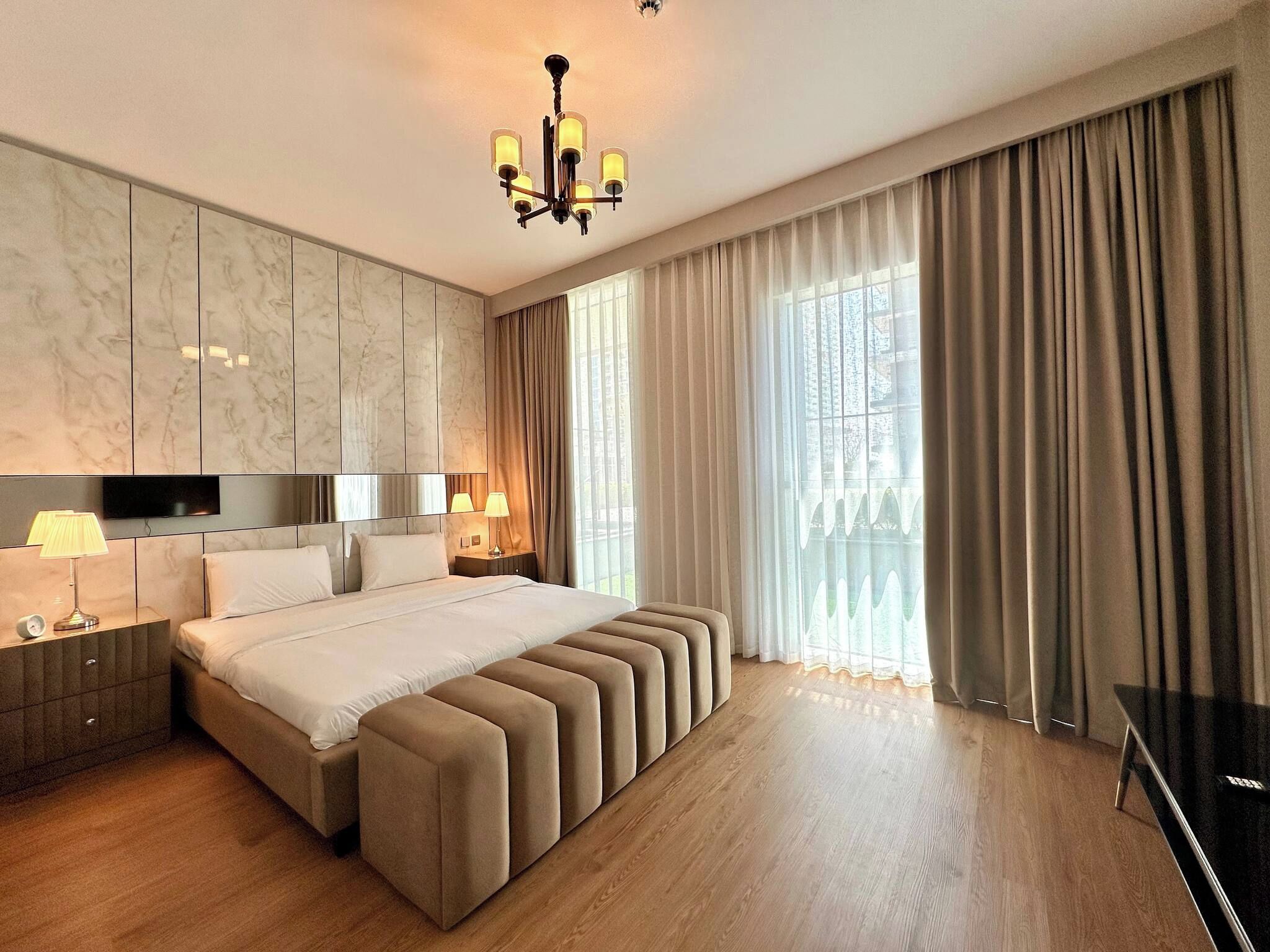 A modern hotel bedroom with a large bed, bedside lamps, a TV, and a chandelier.