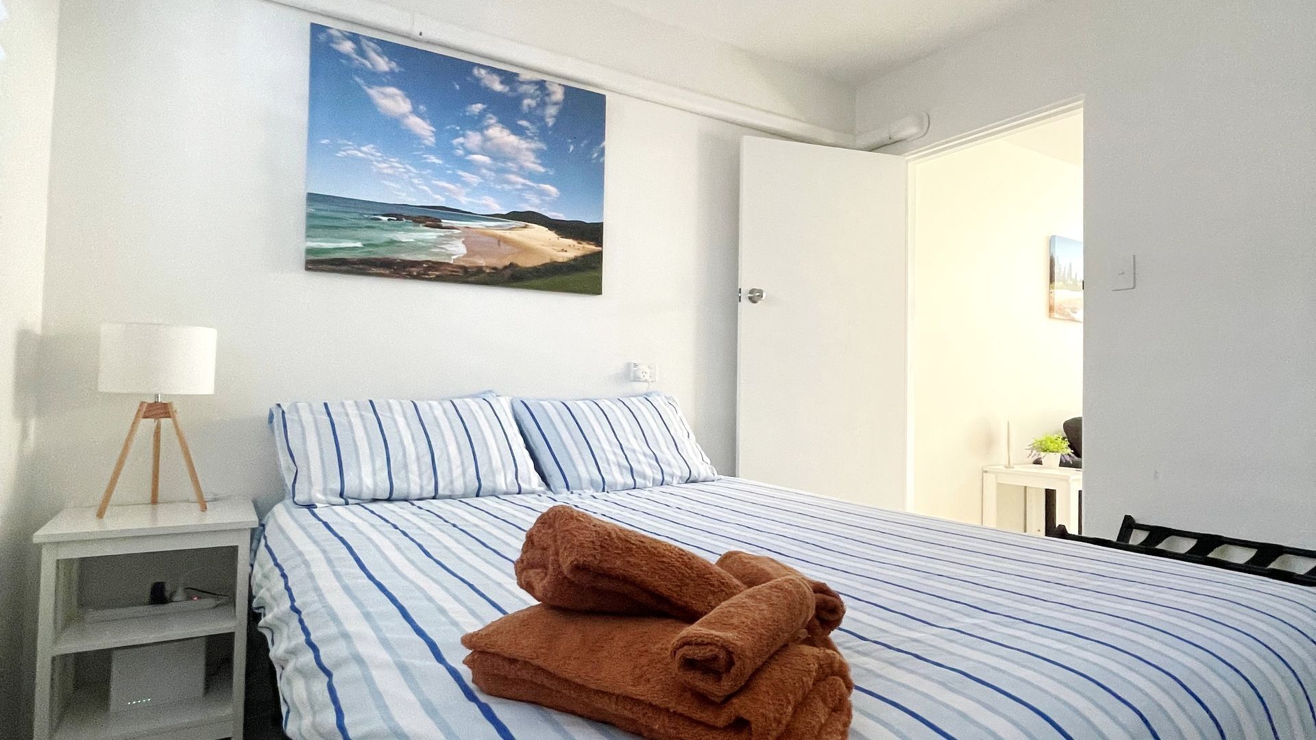 A bedroom with a bed, a nightstand, a lamp, and a painting of a beach scene.