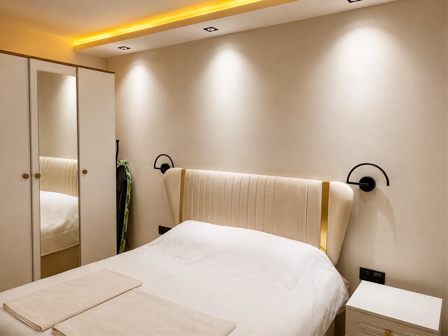 A bedroom with a bed, a nightstand, a bedside table, a mirror, and a wall-mounted light fixture.