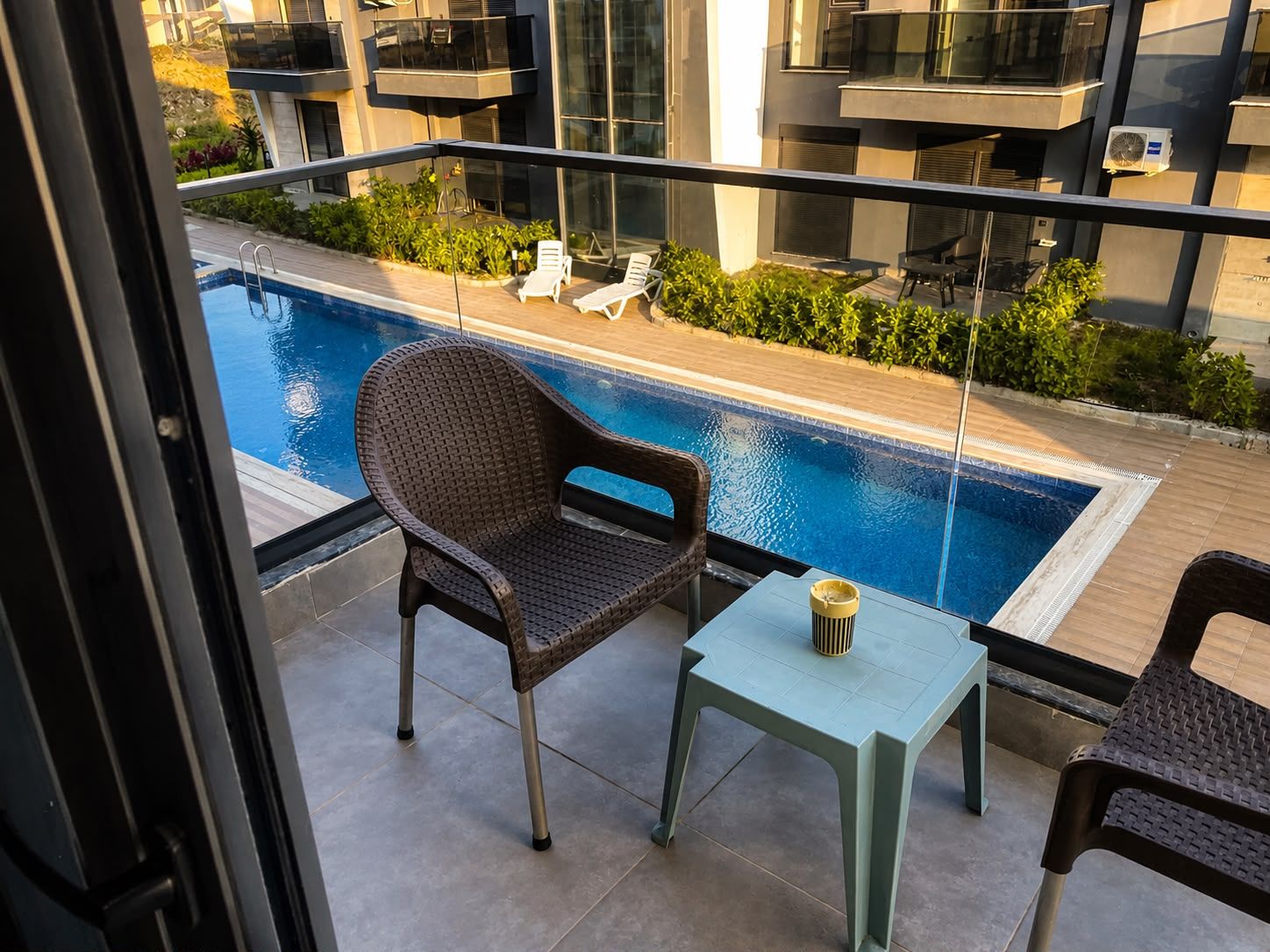 A balcony with a pool, a chair, a table, and a view of a building with balconies.