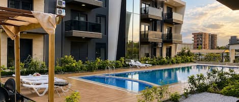 A modern apartment complex with a swimming pool, outdoor seating, and a pergola.