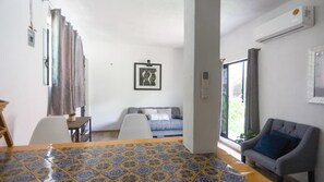 Ein Zimmer mit Esstisch, einer Couch, einem Sessel, einem Fenster mit Vorhängen und einer Klimaanlage.