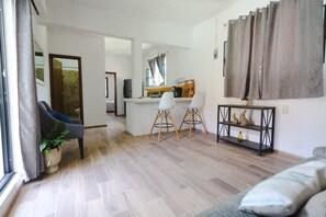 Een woonkamer met een bank, een eethoek met stoelen, een koffieapparaat en een plank met decoratieve objecten.