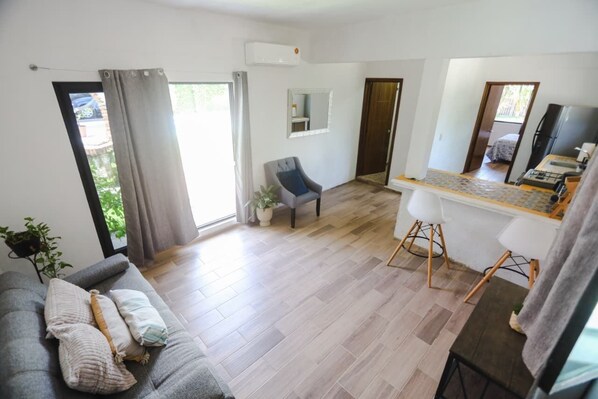 Een moderne woonkamer met een grijze bank, een eethoek met een kleine tafel en stoelen, en een keuken met een koelkast.