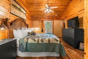Ein Schlafzimmer einer Holzhütte mit einem Bett, einem Schminktisch, einem Deckenventilator und einem Fernseher.