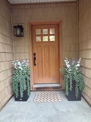 A wooden front door with a glass panel, flanked by two potted plants with white flowers, and a woven doormat on a concrete floor. - Mainland St. Lawrence River Home with Private Dock (Alexandria Bay)