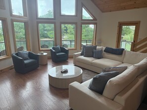 A spacious living room with a high ceiling, large windows, and a view of the outdoors. There are two sofas, a coffee table, and a chair. - Mainland St. Lawrence River Home with Private Dock (Alexandria Bay)