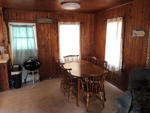 Une pièce aux murs lambrés, avec une table ronde en bois et six chaises, un barbecue et une pancarte indiquant « AU BORD DE LA PLAGE ».