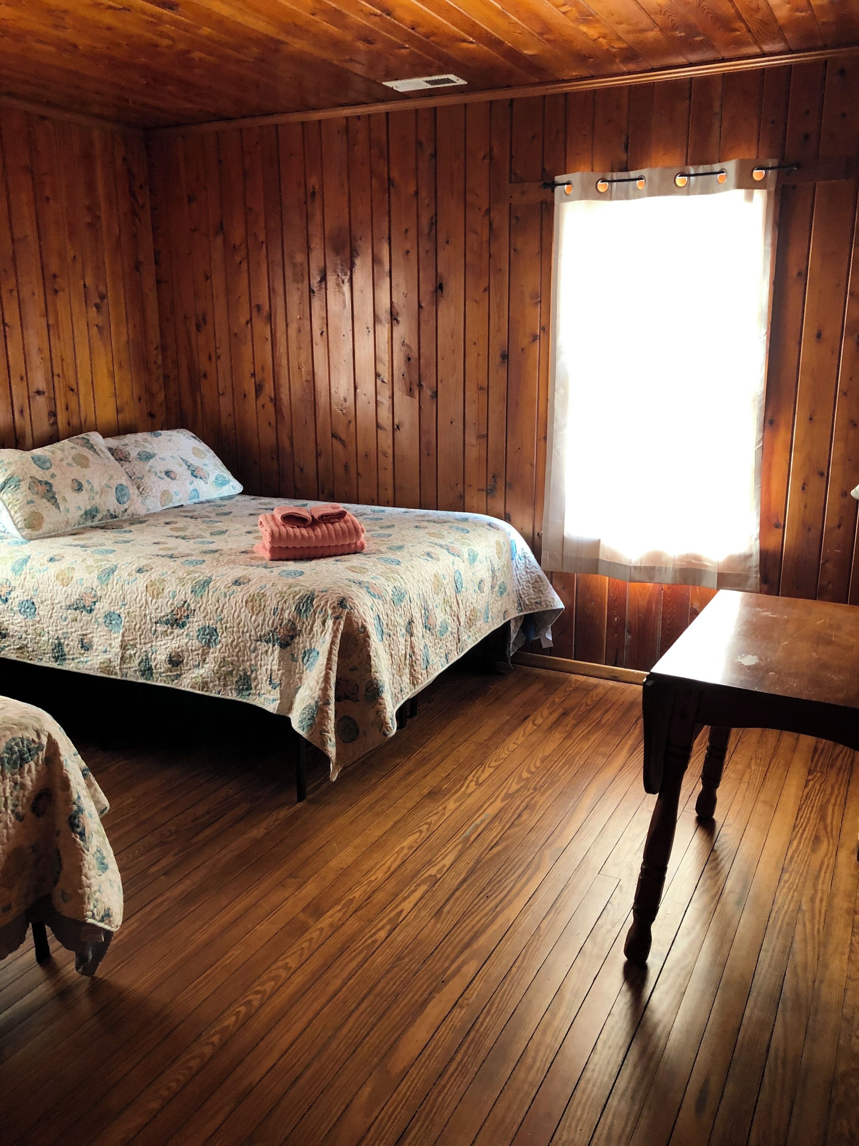 A wooden cabin room with a bed, a table, and a window.