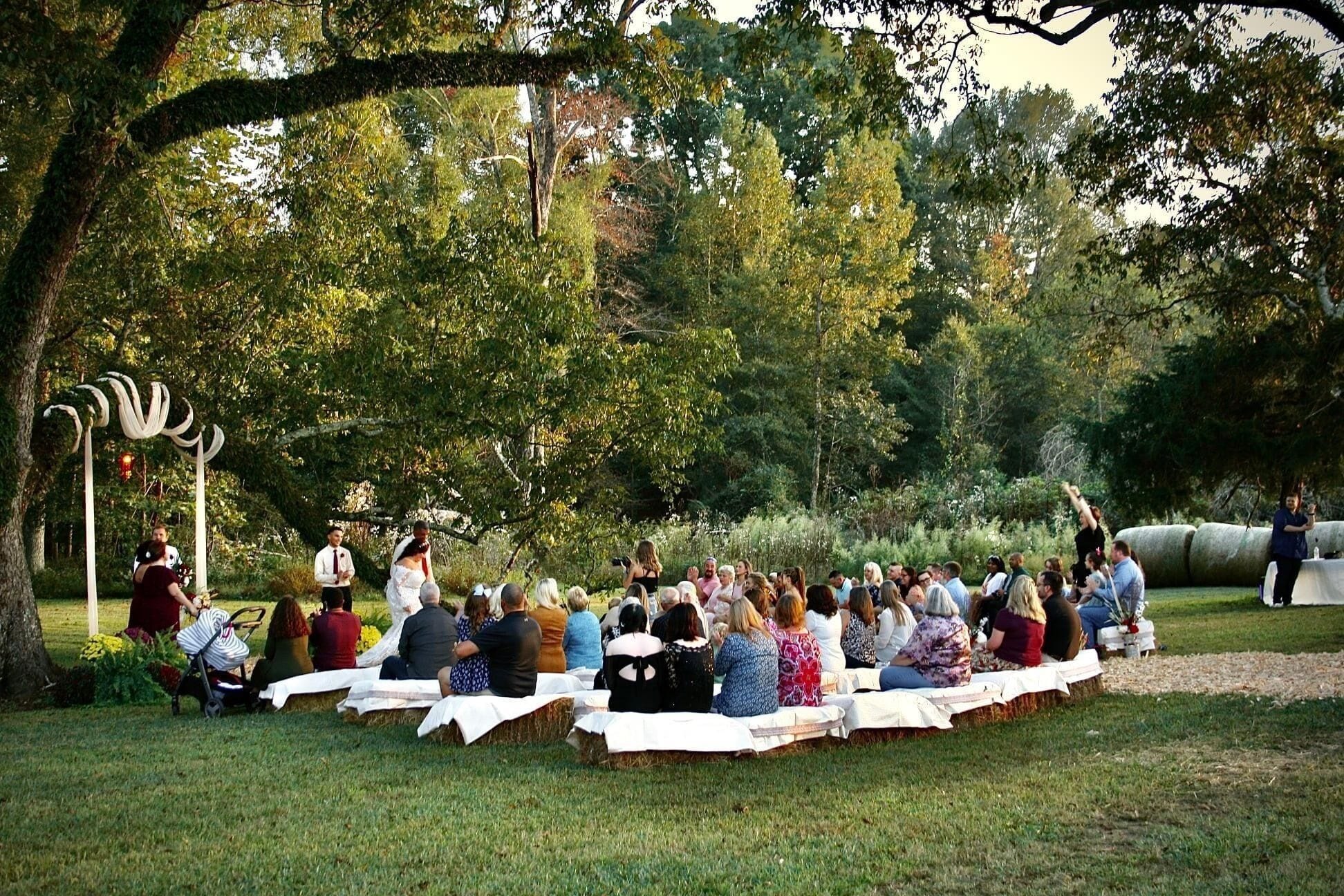 An outdoor gathering with people seated on hay bales under a tree.