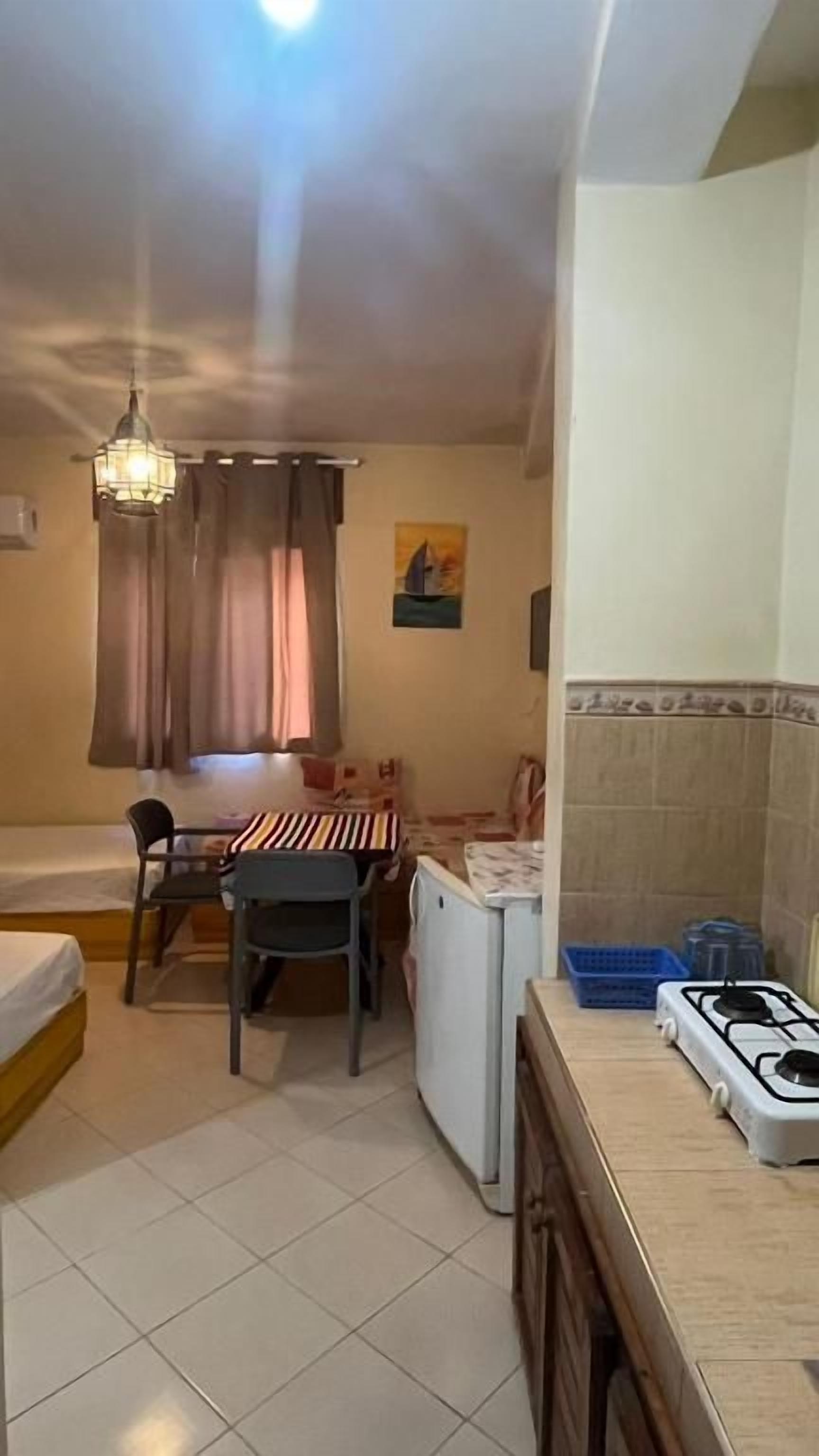 Un espacio habitable compacto con una cama, una mesa de comedor con sillas, una pequeña cocina con estufa a gas y una ventana con cortinas.