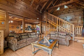Ein Interieur einer Blockhütte mit einer Holztreppe, zwei Sofas mit gemusterten Kissen, einem Holztisch und einem Küchenbereich im Hintergrund.