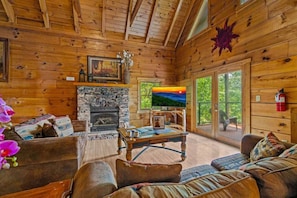 A cozy cabin interior with a stone fireplace, a television, and a large window overlooking greenery. - Epic Views | Hot Tub | Minutes to Pigeon Forge (Sevierville)