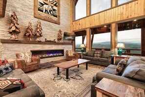 Ein geräumiges Wohnzimmer mit Kamin aus Stein, Holzmöbeln und großen Fenstern mit Blick auf die Berge.