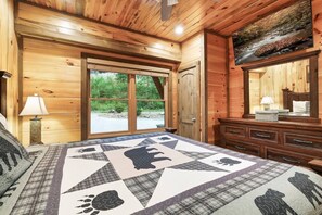 A wooden cabin bedroom with a large bed, a dresser, a mirror, and a window with a view of greenery. - Luxury8bdrmCabin-LgPool-HotTub-5MinParkway-Special (Sevierville)