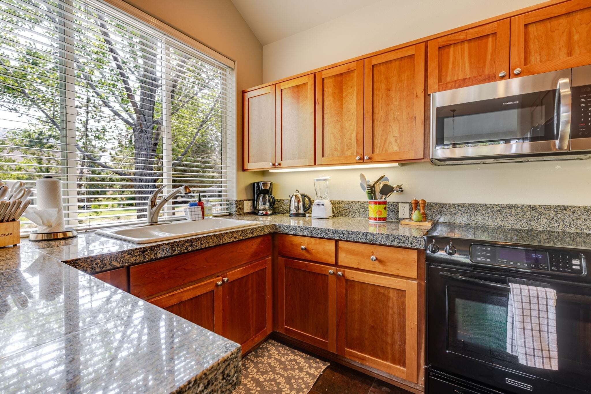 A kitchen with wooden cabinets, a stainless steel microwave, a stove, and a window with blinds.