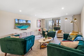 A modern living room with a pool view, green sofas, a flat-screen TV, and a large window. - Desert Elegance Off El Paseo (Palm Desert)
