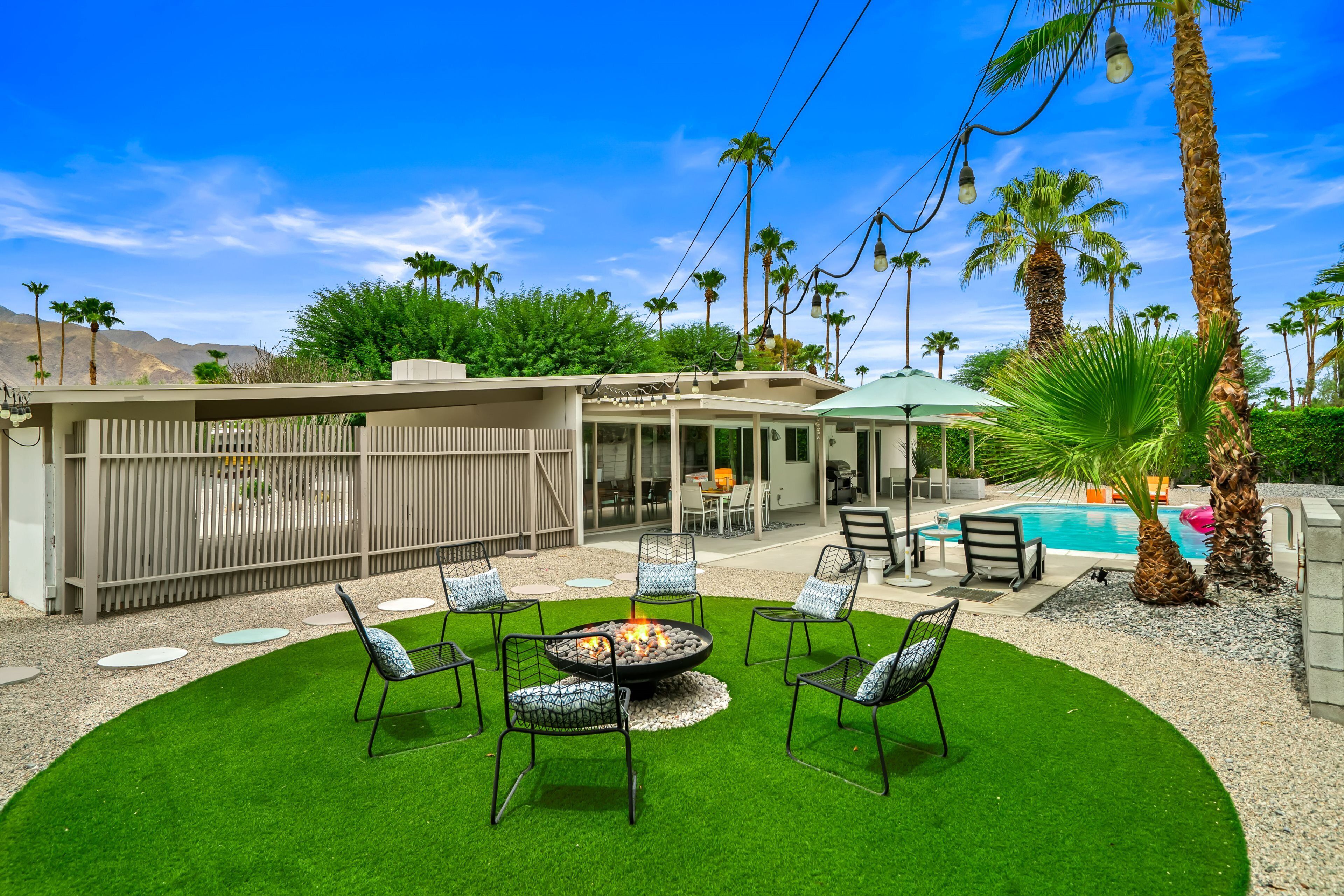 A backyard with a fire pit, outdoor seating, a pool, and palm trees.