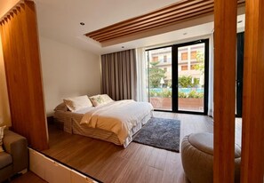 Een moderne slaapkamer met een groot bed, een kleine zithoek en uitzicht op een zwembad door een glazen schuifdeur.