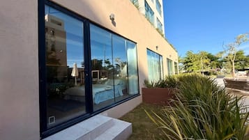 Un bâtiment moderne doté de grandes fenêtres, dont l’intérieur est visible et comprend un lit et un coin repas, ainsi qu’un jardin bien entretenu avec des plantes.