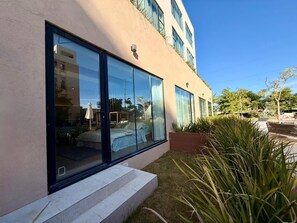 Un bâtiment moderne doté de grandes fenêtres, dont l’intérieur est visible et comprend un lit et un coin repas, ainsi qu’un jardin bien entretenu avec des plantes.