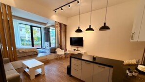 En moderne stue med sofa, sofabord og fjernsyn. Der er et køkkenområde med skabe og en bordplade.