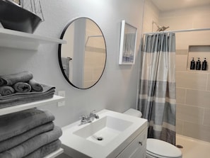 A bathroom with a white sink, mirror, and toilet. There are towels stacked on a shelf, and a shower curtain with a striped pattern. - Peaceful 2 Bedroom apartment Near Denton Medical Center (Denton)