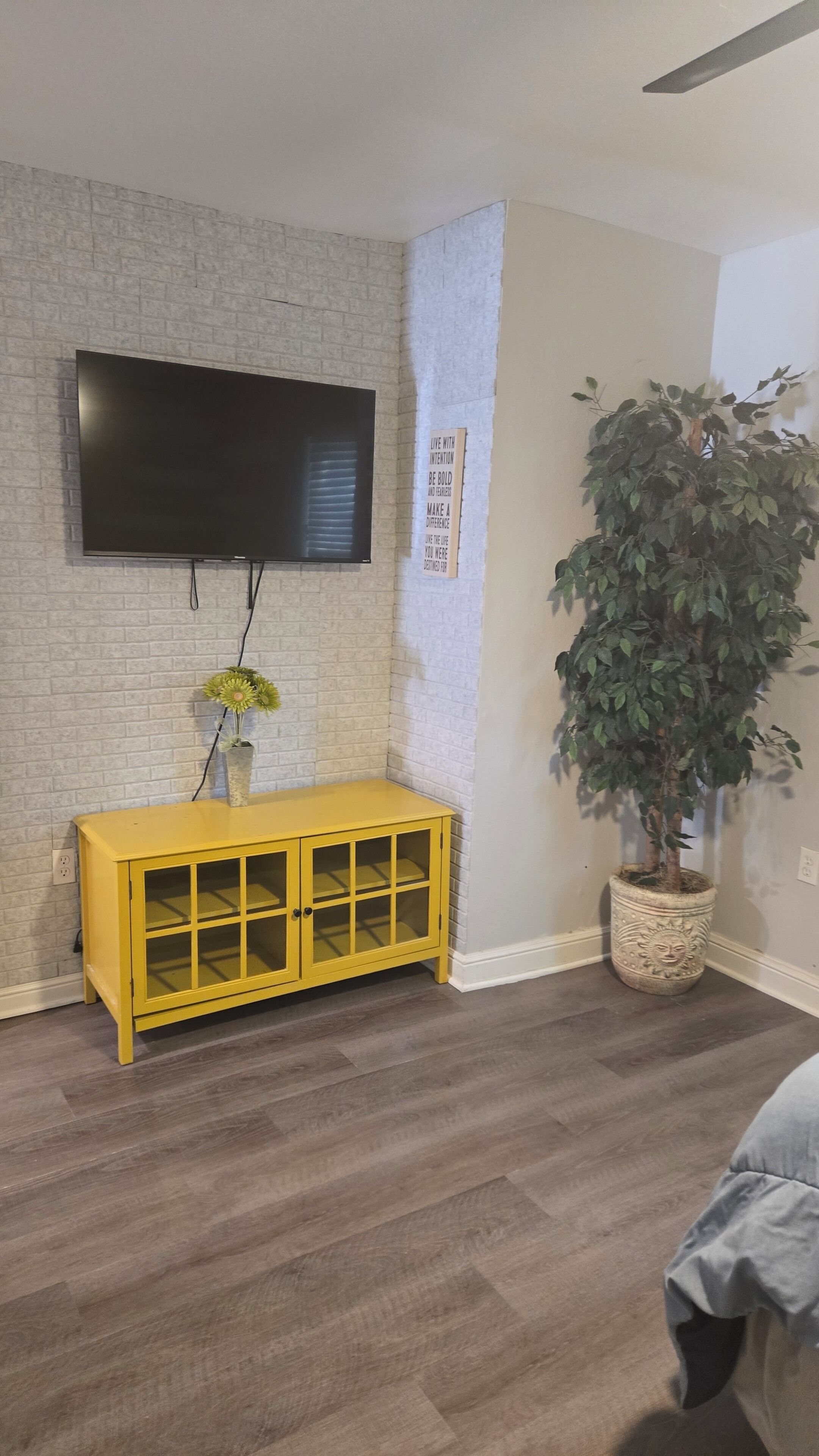 A room with a flat-screen TV mounted on a brick wall, a yellow console table with glass doors, a potted plant, and a ceiling fan.