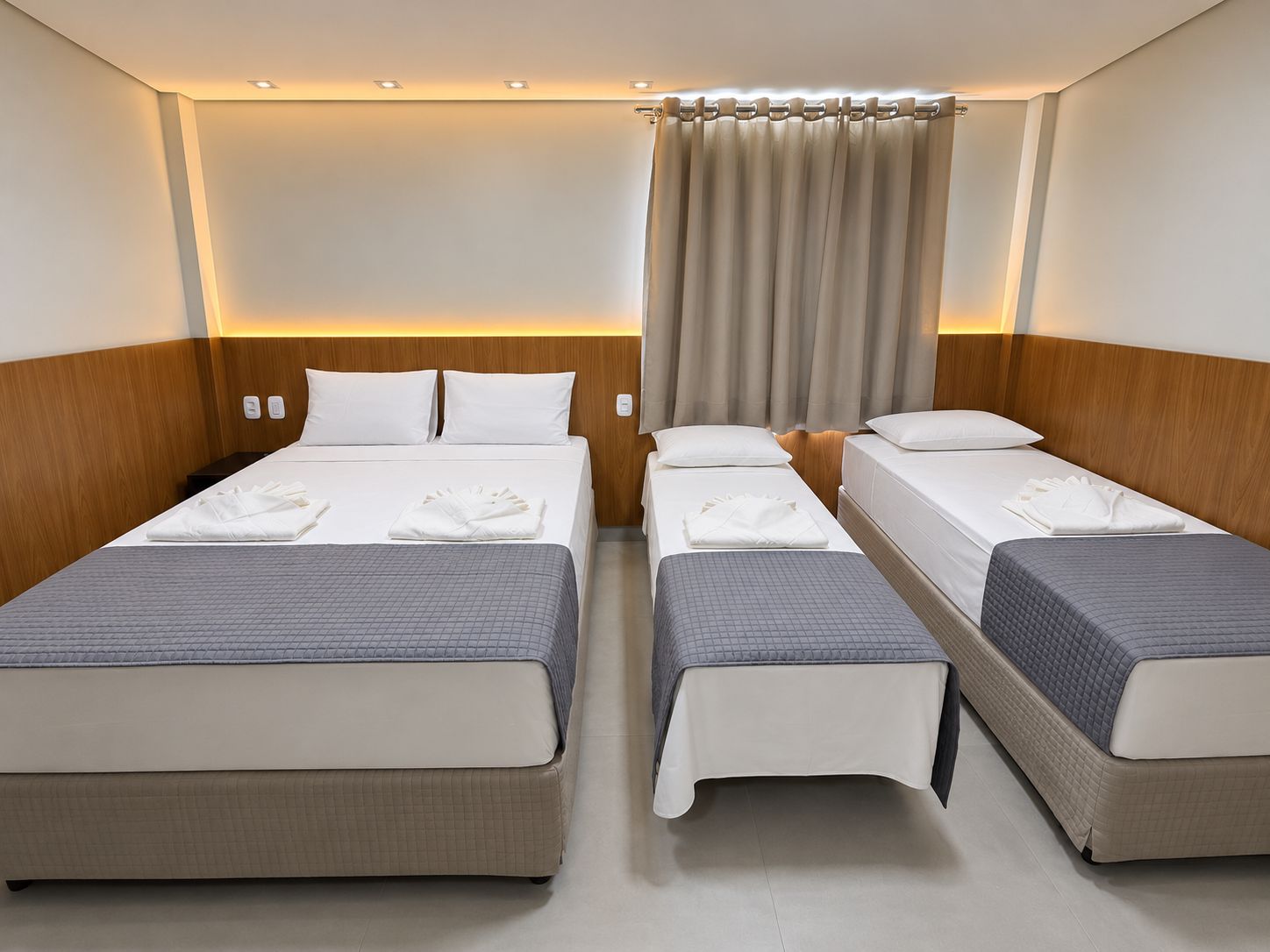 Une chambre d’hôtel avec deux lits simples, une tête de lit en bois, un rideau et une table de chevet.