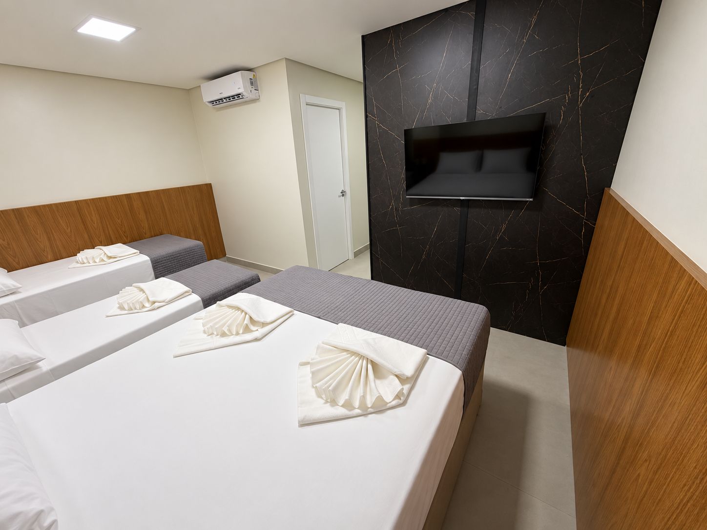 Habitación de hotel con tres camas, un televisor de pantalla plana montado en una pared de mármol oscuro y un equipo de aire acondicionado instalado en el techo.