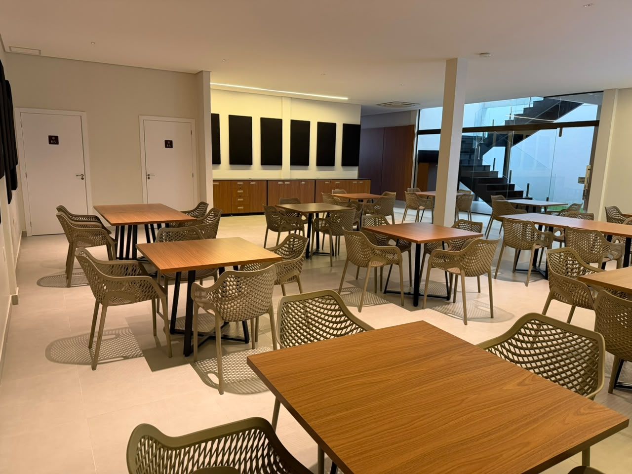 Un comedor moderno con varias mesas y sillas de mimbre, una escalera al fondo y vistas al exterior.