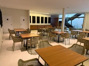A modern dining area with multiple tables and wicker chairs, a staircase in the background, and a view of the outdoors.
