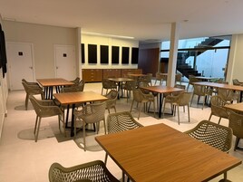 Uma sala de jantar moderna com várias mesas e cadeiras de vime, uma escada ao fundo e vista para o exterior.