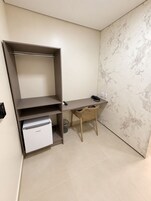 Quarto compacto com escrivaninha, cadeira, eletrodoméstico pequeno branco e prateleira na parede.