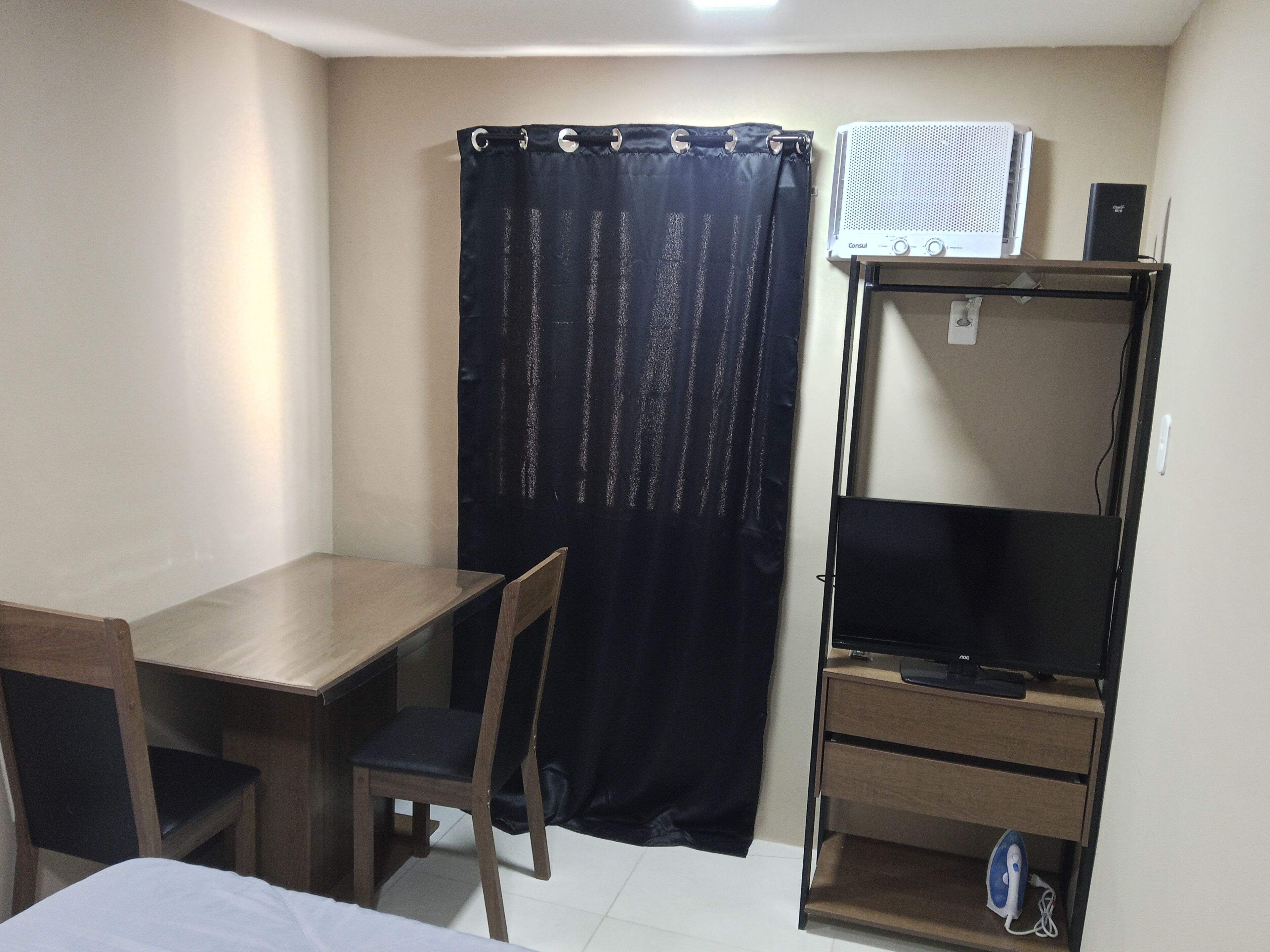 Quarto pequeno com cama, escrivaninha, cadeira, televisão sobre suporte e cortina.