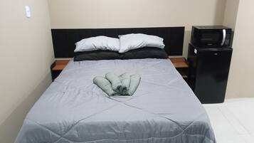 Una cama con una colcha gris, dos almohadas blancas y una toalla doblada. A la derecha de la cama, un mueble negro para televisión con un microondas.