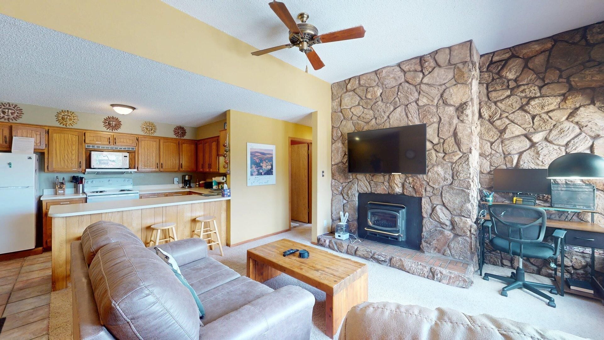 A living room with a sofa, coffee table, and a fireplace. There is a kitchen area with wooden cabinets and a microwave. A ceiling fan is visible, and a television is mounted on the wall.