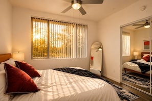 A bedroom with a bed, window blinds, a ceiling fan, a bedside lamp, and a mirror. - Bright Tempe Studio • Green Views • 5 Min to A (Tempe)